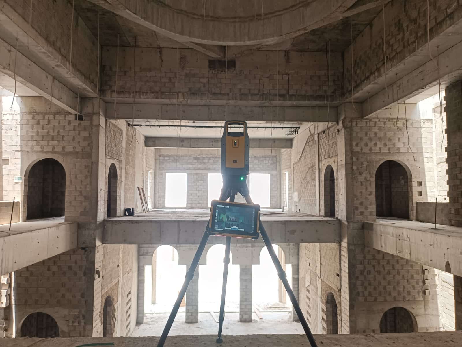 A 3D laser scanner mounted on a tripod with a connected tablet controller, performing a digital scan inside a multi-story concrete building under construction.
