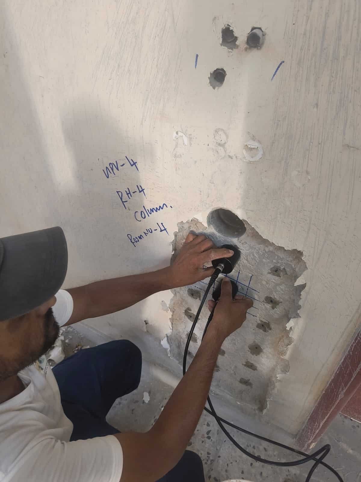 An engineering technician performing non-destructive concrete testing (NDT) using an ultrasonic pulse velocity transducer on an exposed concrete structural column labeled Room No. 4.